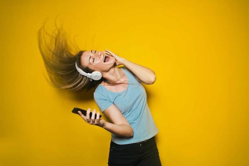 Woman wearing headphones and holding a smartphone is flicking her hair while dancing with her eyes closed.