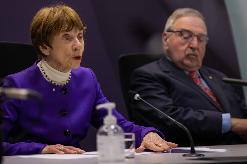 A woman with short dark hair and a purple jacket sits at a table in front of a microphone next to a man with a moustache..