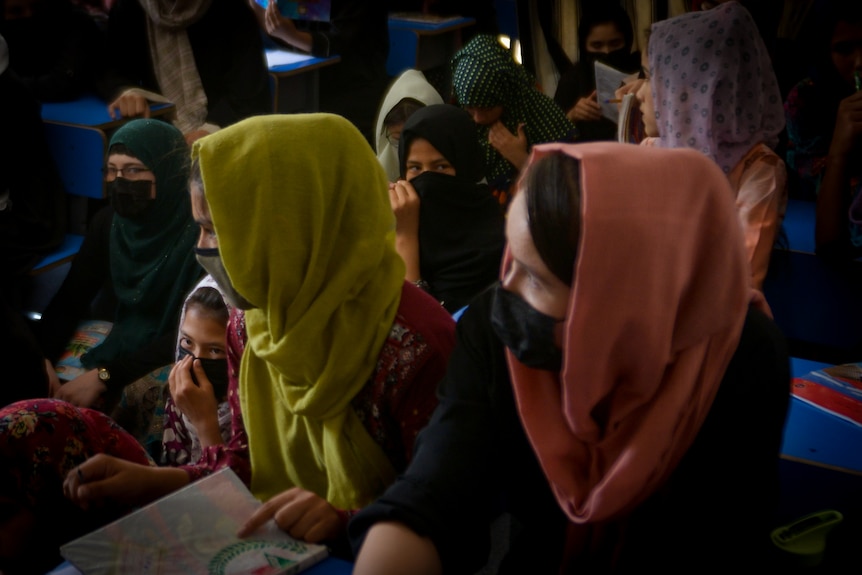 Girls in colourful headscarves sit at rows of desks and on the ground in a small dark room.