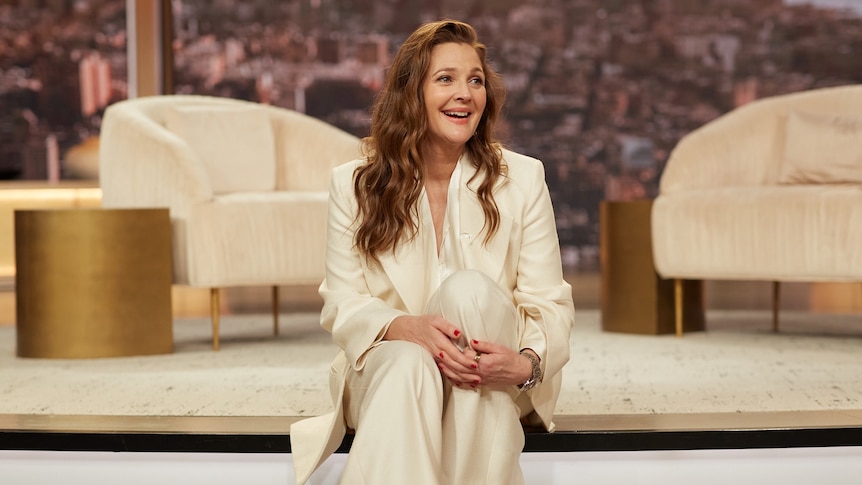 A white woman with long hair wears a flowing suit and sits on a stair and smiles, on the set of a daytime TV talk show