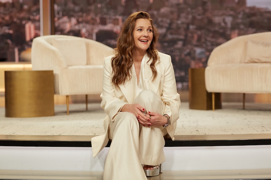 A white woman with long hair wears a flowing suit and sits on a stair and smiles, on the set of a daytime TV talk show