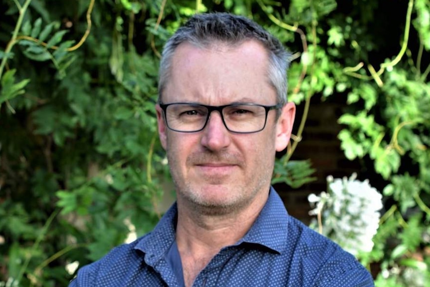 Jason Thompson, wearing a blue shirt and glasses, stands in a garden.