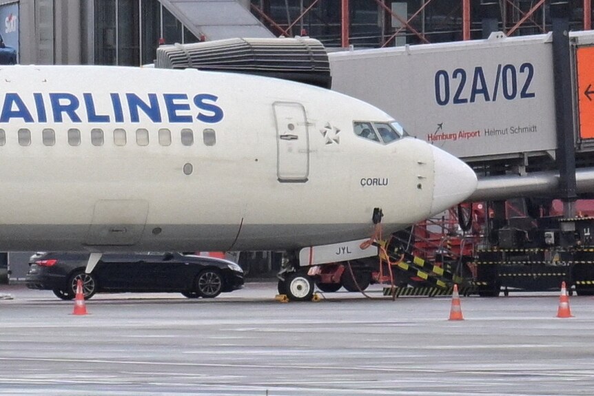 An image of the nose of an airplane, behind it is a parked car.