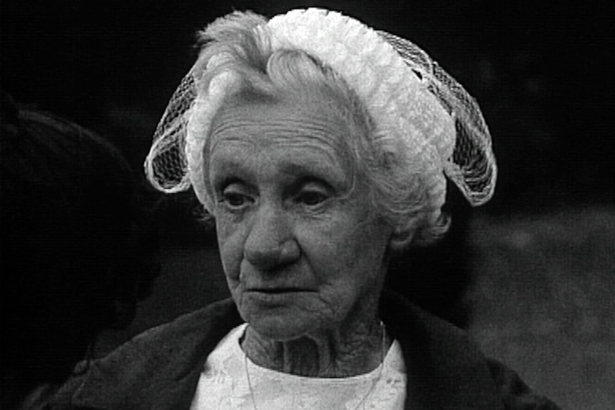 A historical black-and-white image of an elderly woman standing in front of the front brick walls of a jail. She looks frail