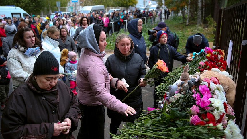 People, mostly women in this photo, cry and lay pink and orange flowers at a school gate.