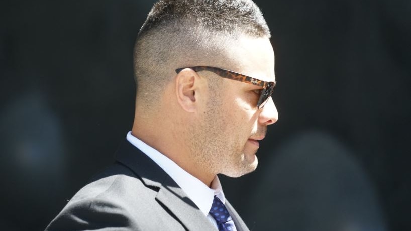 A man with short hair, wearing dark sunglasses with tortoiseshell frames, stands in profile, the sun hitting his face.