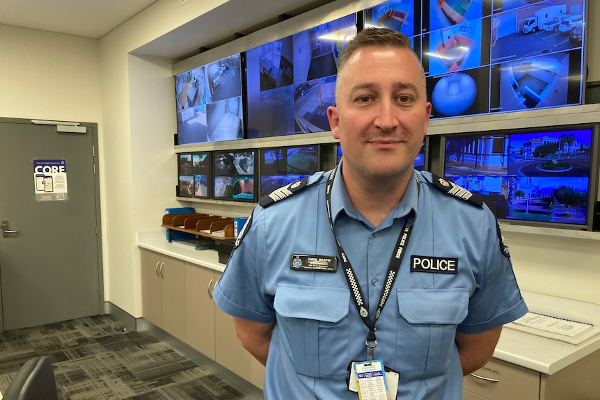 Smiling police officer, in blue uniform, standing in front of wall of security cameras.