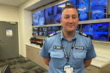Smiling police officer, in blue uniform, standing in front of wall of security cameras.