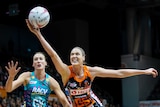 A netball player stretches for the ball while a defender runs up behind her.