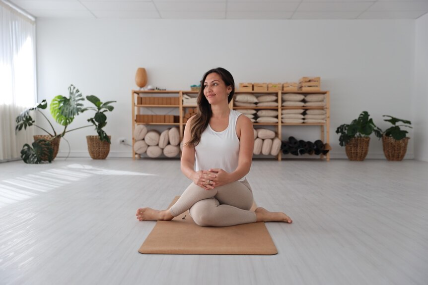 Bright white room with green plants and yoga props in background, woman with brown hair practicing sun salutations on brown mat