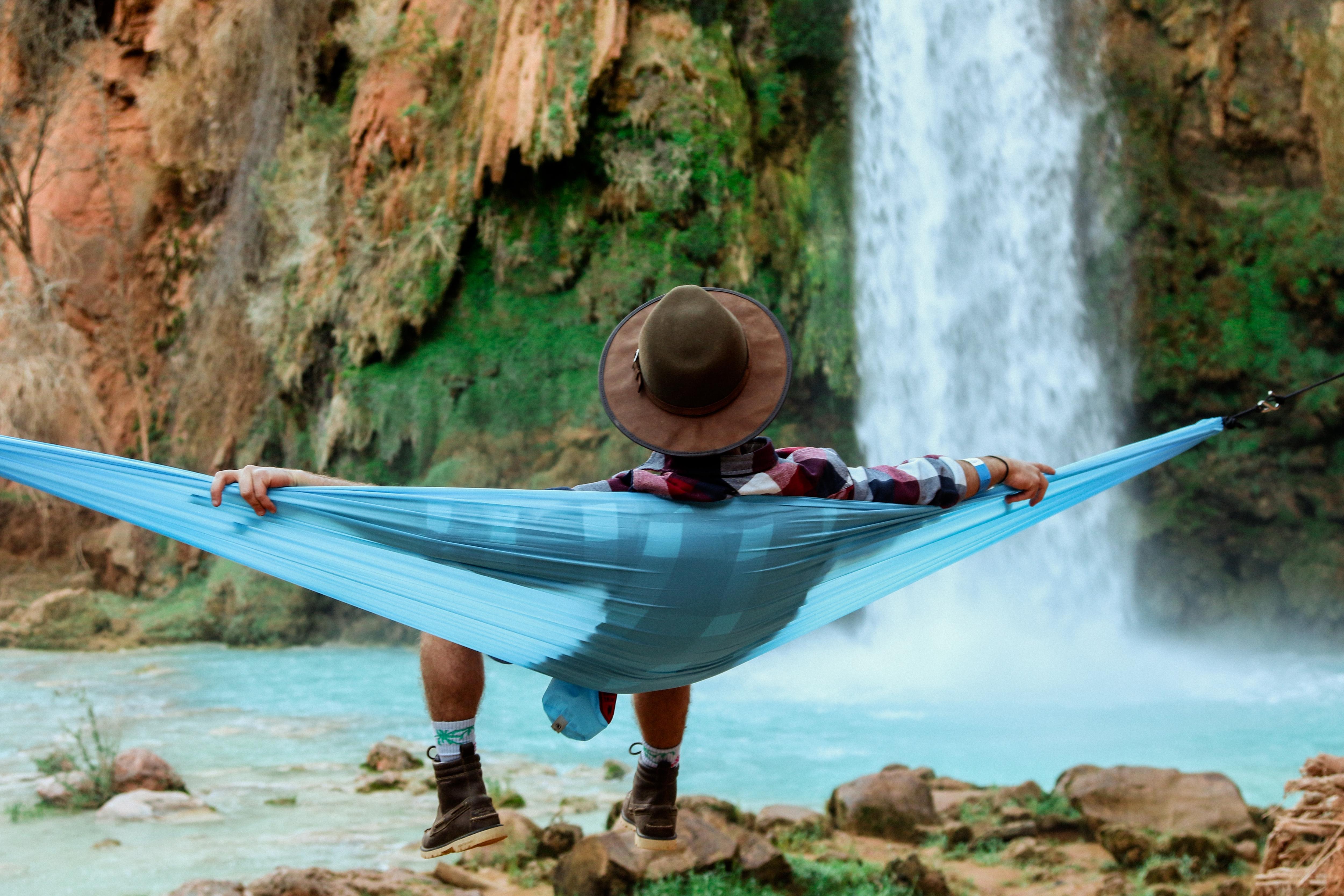 An image from behind of a man relaxing in a hammock near a waterfall. He is wearing a wide-brimmed hat.