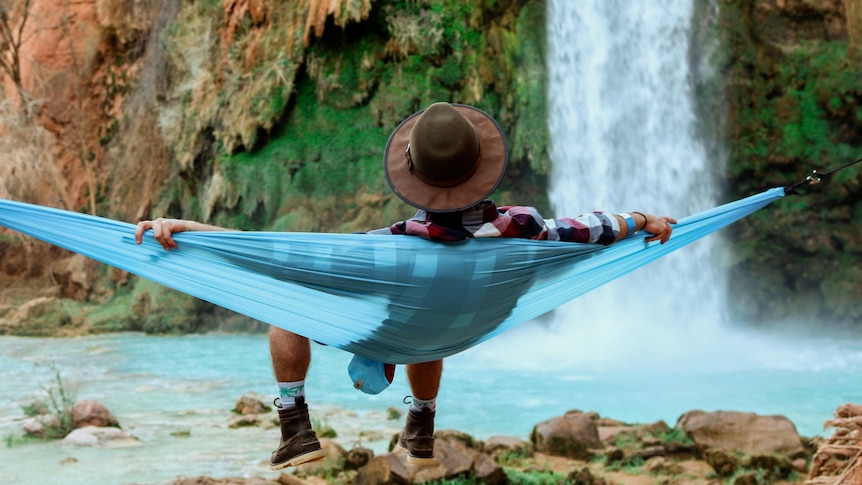 An image from behind of a man relaxing in a hammock near a waterfall. He is wearing a wide-brimmed hat.