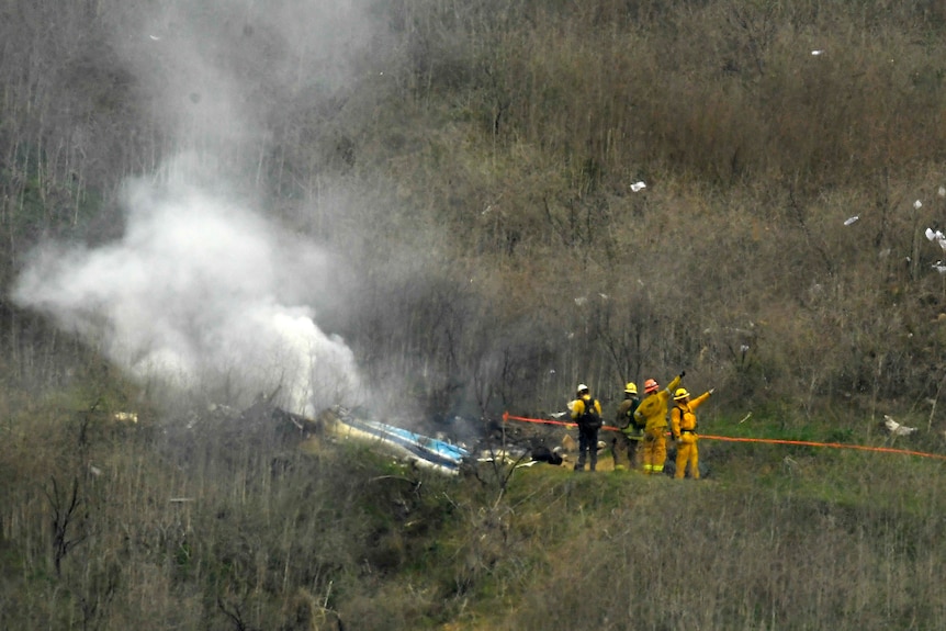 A zoomed-in photo shows firefighters in yellow hi-vis uniforms working next to smoking wreckage on a tree-lined hillside.