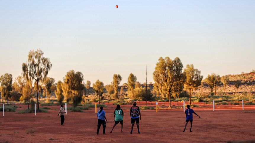 Young Aboriginal women kick a football on red dirt in the early evening light 