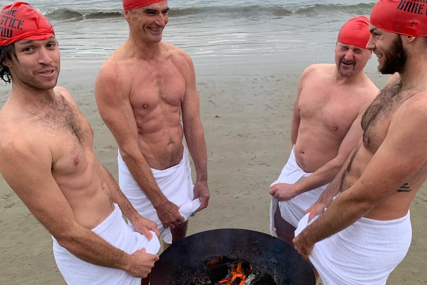 Four naked men wrapped in towels wearing red swim caps warm their nether regions over a fire-pot after a mid-winter nude swim