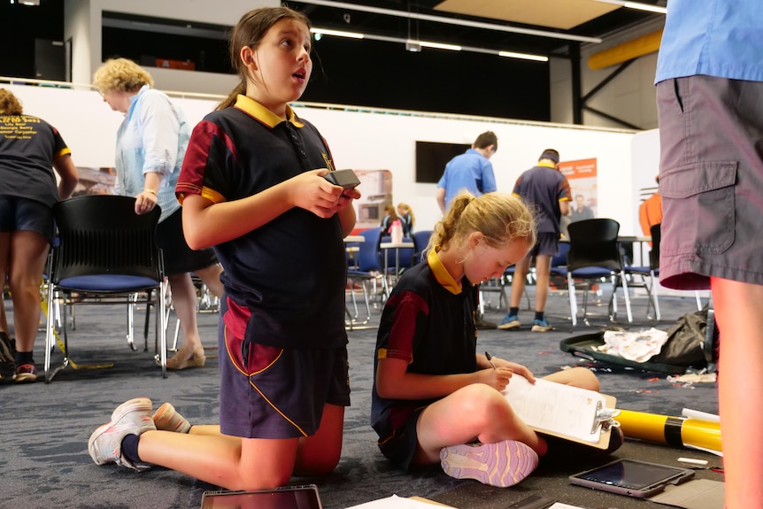 Two students, one on their knees reading a clue, the other sitting crossed legged with a clipboard.