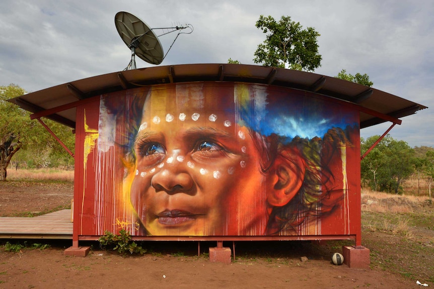 The photograph of the outside of a demountable school building with a curved tin roof and a mural of an Aboriginal boy on wall.