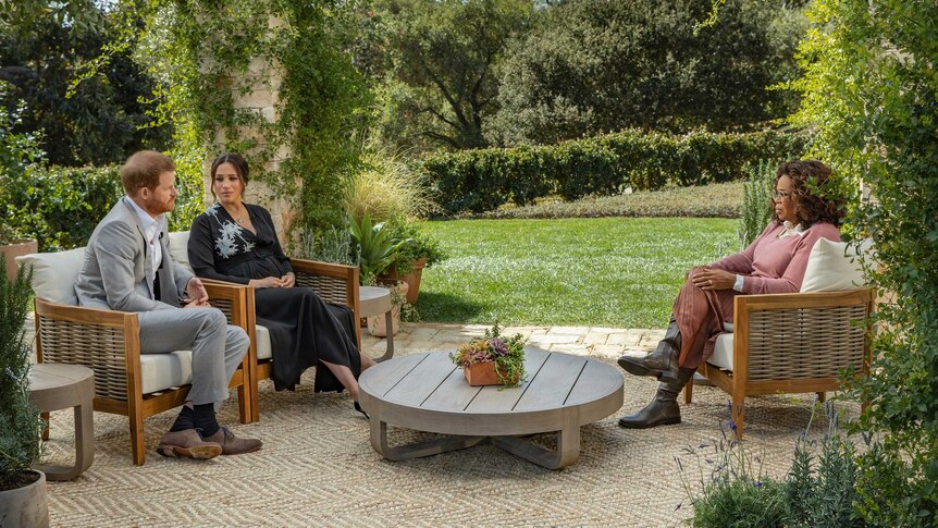 Prince Harry and Meghan sit on armchairs opposite Oprah Winfrey on a patio in a lush, green garden.