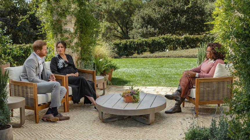 Prince Harry and Meghan sit on armchairs opposite Oprah Winfrey on a patio in a lush, green garden.