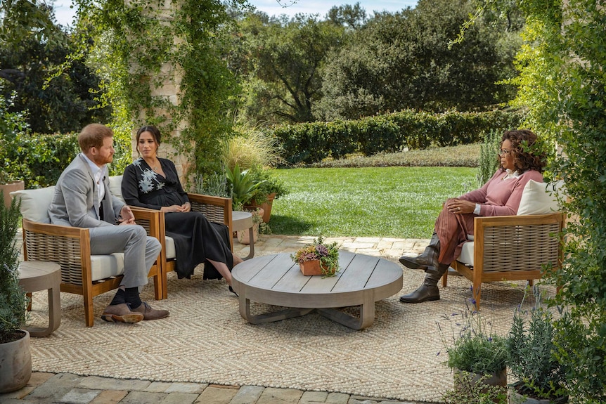 Prince Harry and Meghan sit on armchairs opposite Oprah Winfrey on a patio in a lush, green garden.