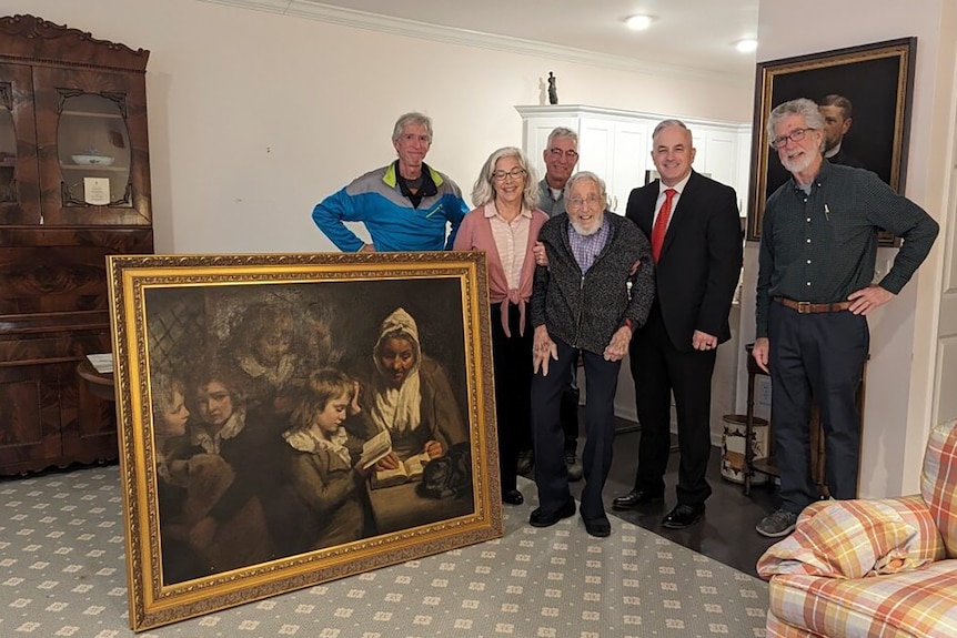 Group of people stand around gold framed painting in home. 