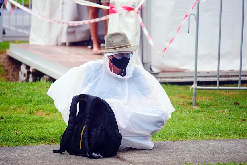 A man, wearing a white raincoat, sits on a footpath outside a COVID testing clinic