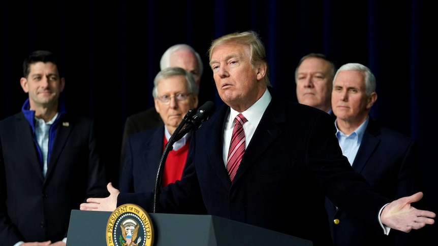 Donald Trump speaks at a podium with his colleagues standing around him.