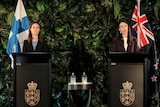 Two women stand at lecterns on a stage. One has the Finland flag behind her, and the other has the New Zealdn flag behind her.