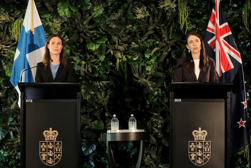 Two women stand at lecterns on a stage. One has the Finland flag behind her, and the other has the New Zealdn flag behind her.