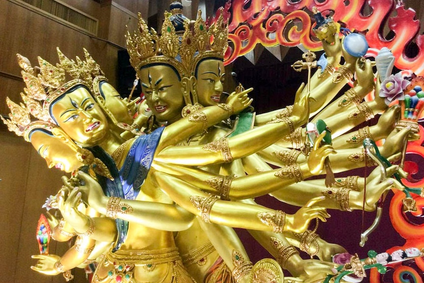 A gold coloured statue of two beings, each with four faces an multiple arms, in sexual union while standing up.