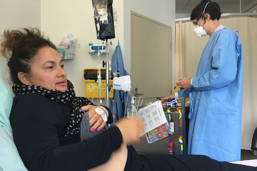 A woman in a hospital chair raises her shirt as she is administered drugs via a drip and tended to by a person in scrubs.