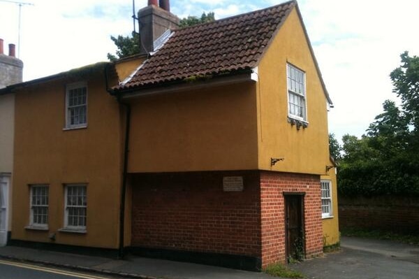 Street view of cottage with triangle roof and tiles, mustard yellow facade on top of brick bottom level.