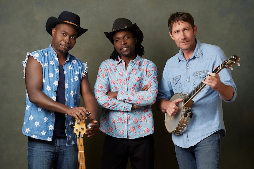 Two black men, one white carrying banjos, holding a guitar, two wearing cowboy hats