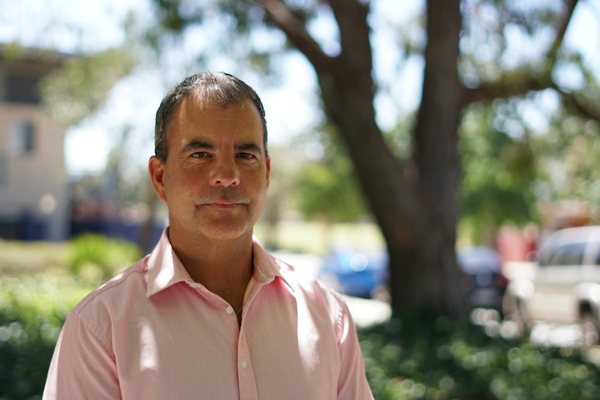 A man with short, dark hair stands in dappled light beneath a tree.