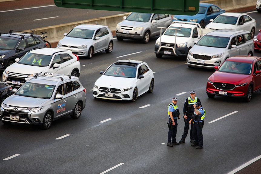 A group of three police officers standing in front of stationary cars.