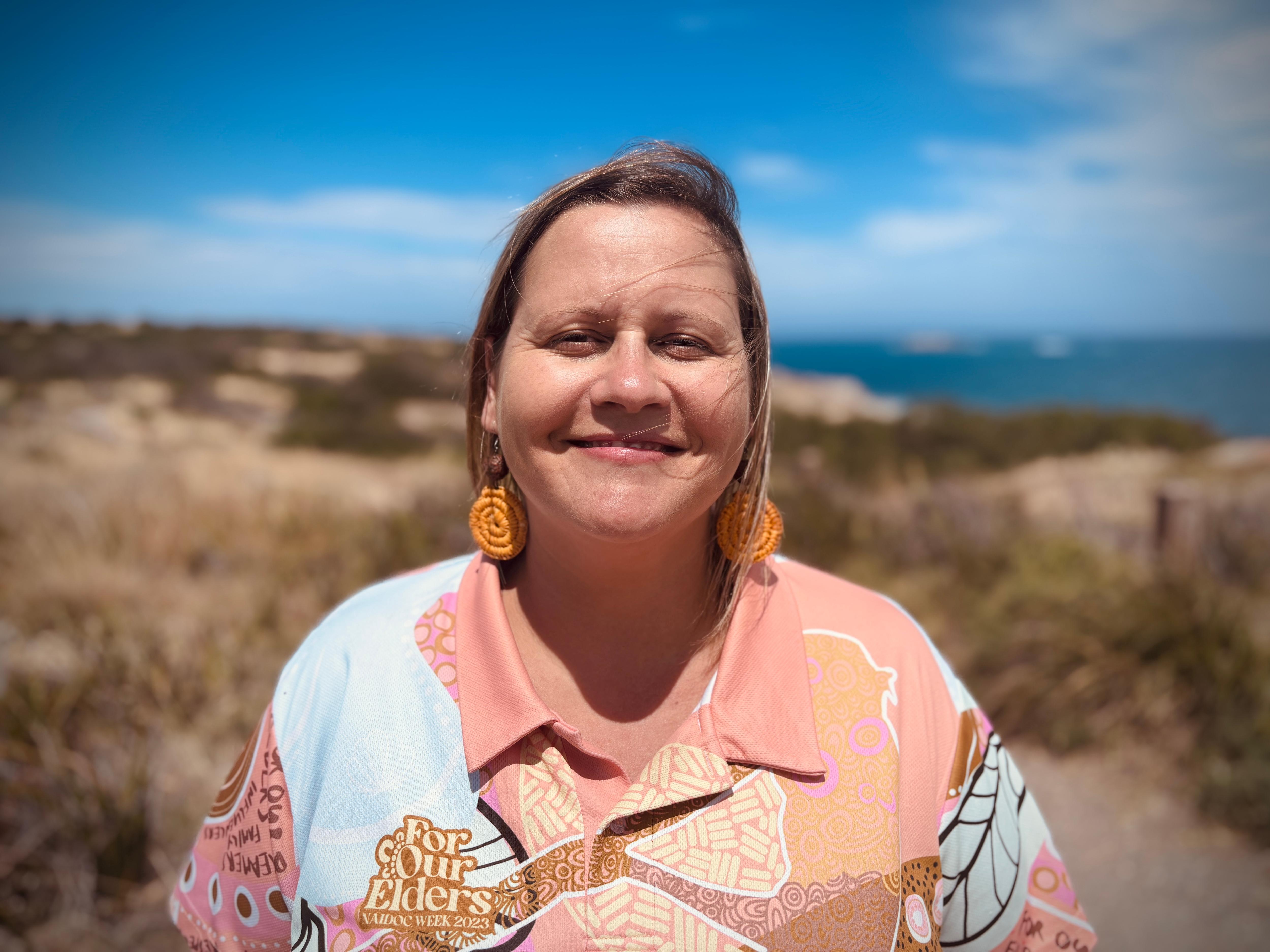 Una mujer con cabello castaño rubio que sopla sobre su rostro con el viento sonríe y usa aretes trenzados. El mar está al fondo.