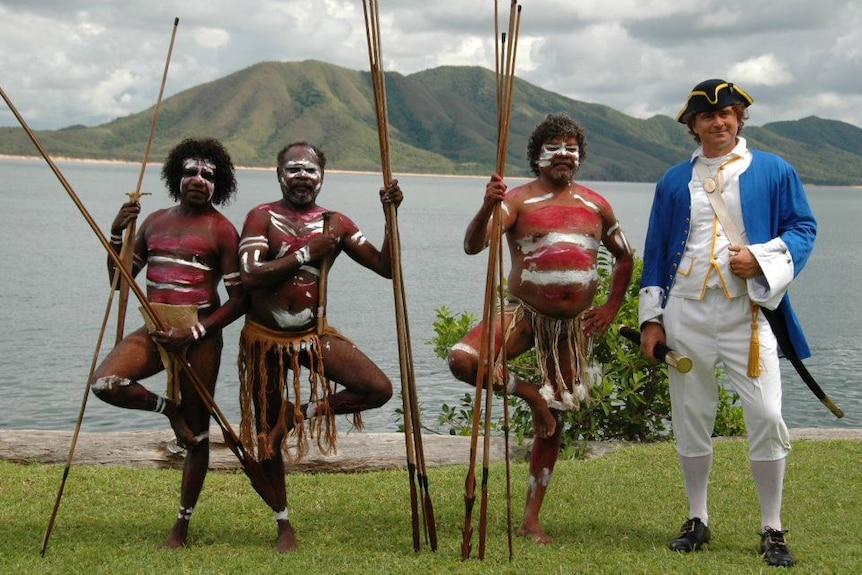 A man dressed in clothes from 1770 stands with three aborigines wearing traditional body paint on a shore.