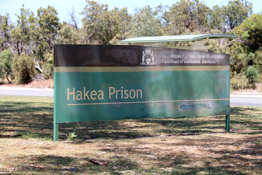 A red and black Hakea Prison sign on grass beside a road.