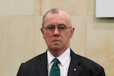 Richard Tracey, wearing a dark suit, stands in front of a sign for the aged care royal commission.