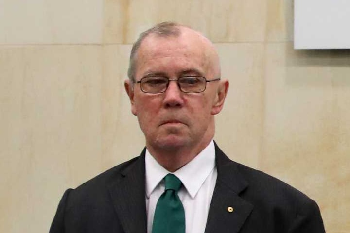 Richard Tracey, wearing a dark suit, stands in front of a sign for the aged care royal commission.