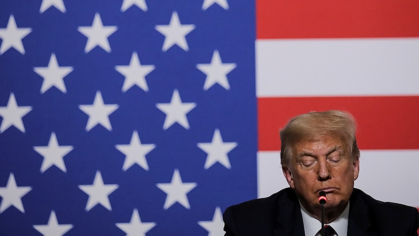 Donald Trump looks downcast in front of a large US flag.