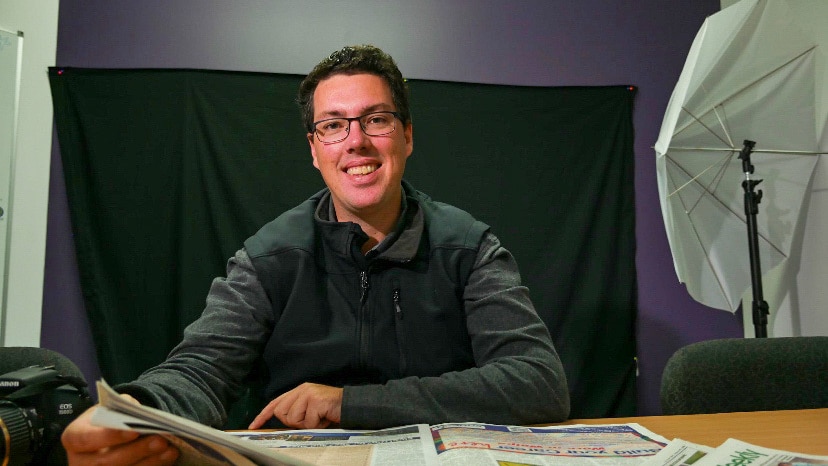 Man sits smiling at camera with a newspaper in hand. 