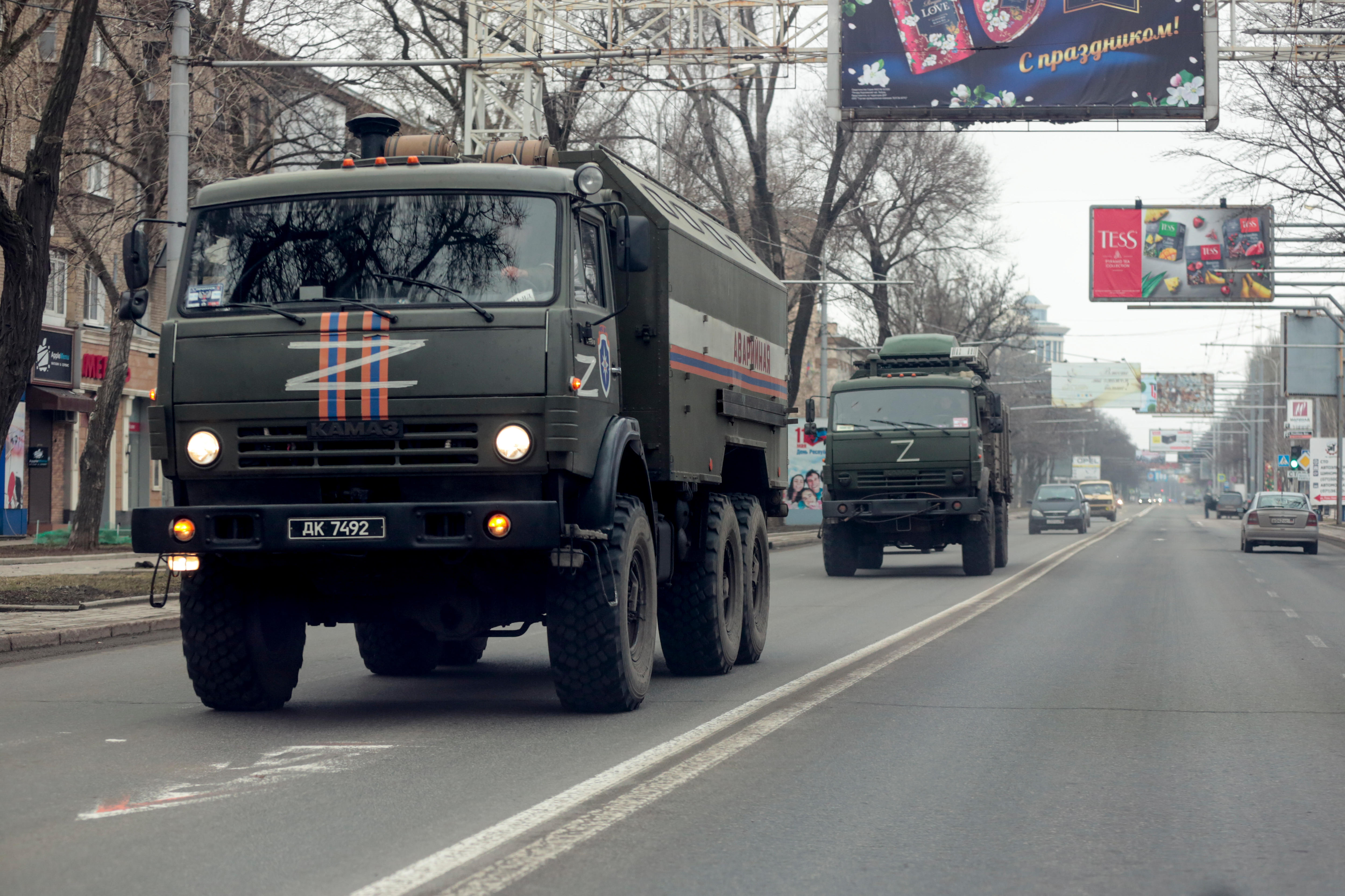Two large military trucks drive down a road, with civilian cars in the background.
