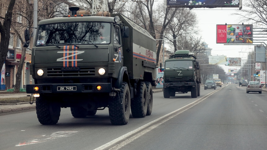 Two large military trucks drive down a road, with civilian cars in the background.