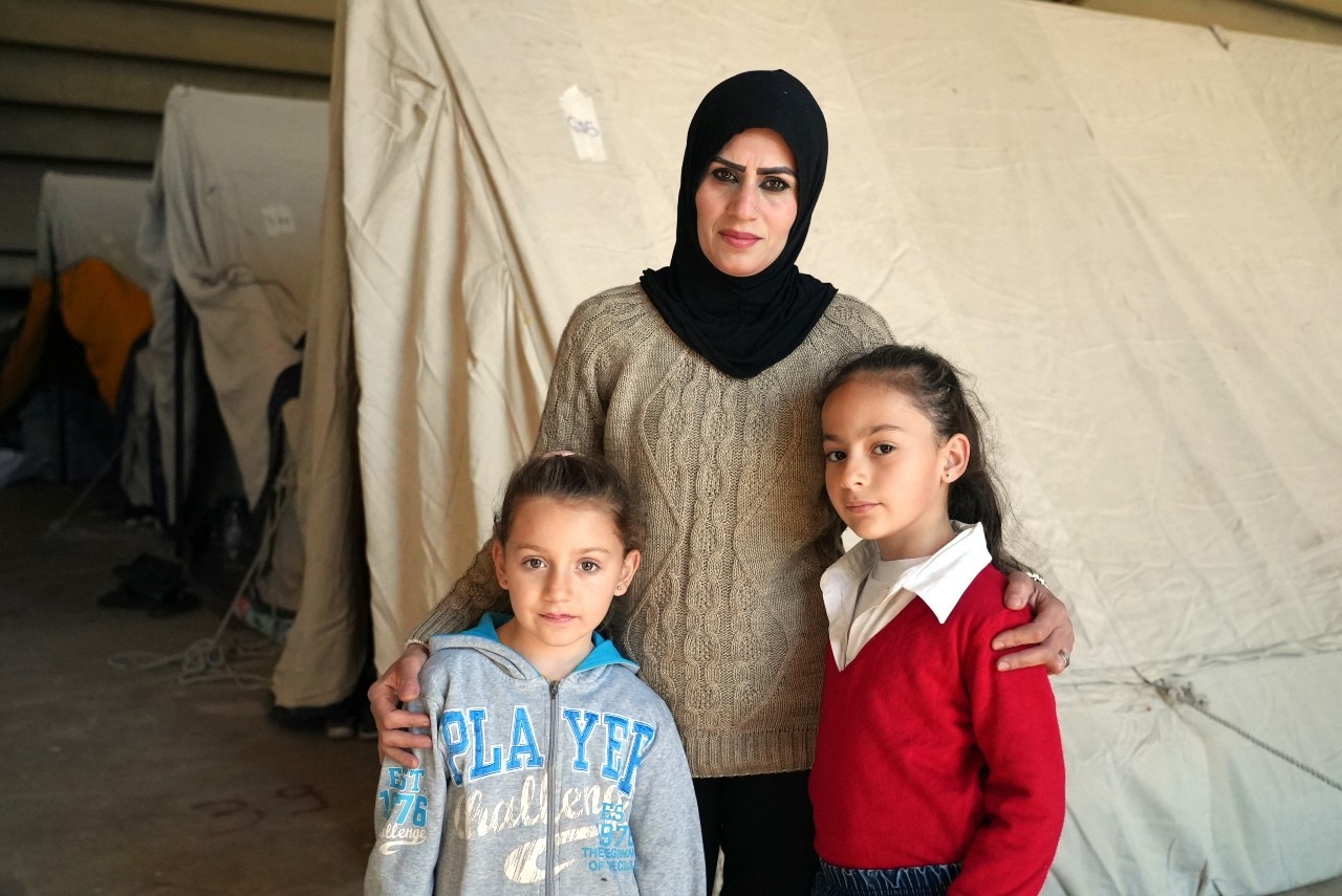 A woman looks at the camera with a neutral experssion. She has her arms around two young children.