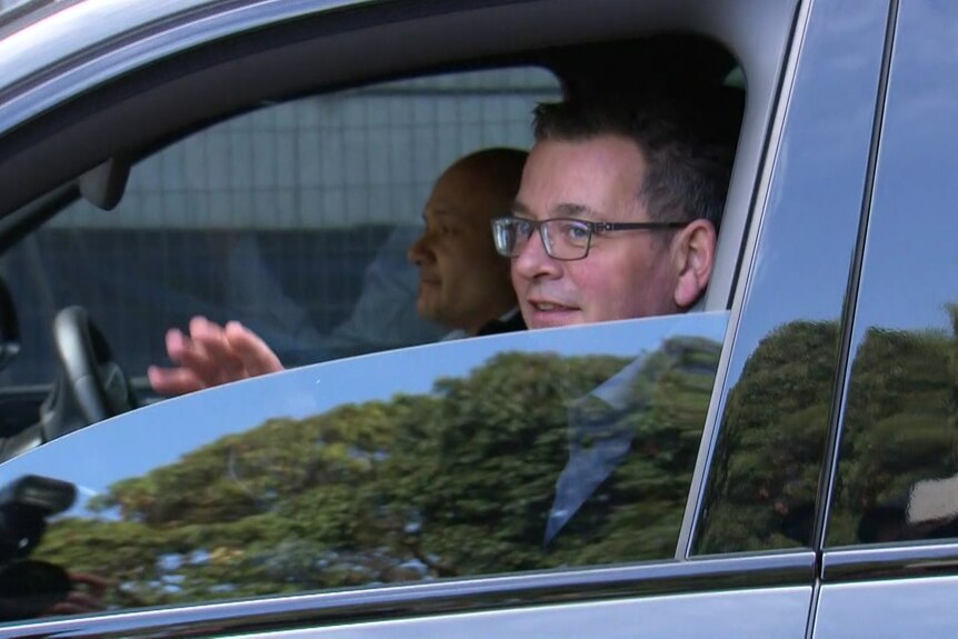 Andrews sits in the front passenger seat of a white car and waves out the half open window.