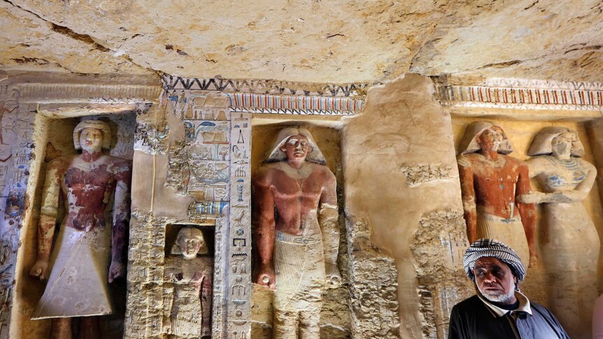 An excavation stands in front of a wall that has ancient Egyptian statues and hieroglyphs carved in.