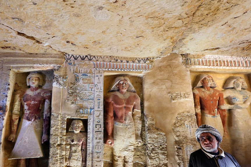An excavation stands in front of a wall that has ancient Egyptian statues and hieroglyphs carved in.