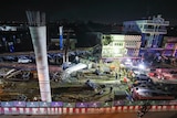 An elevated view of a construction site at night. It is lit up with spotlights. Debris is strewn around and workers can be seen.