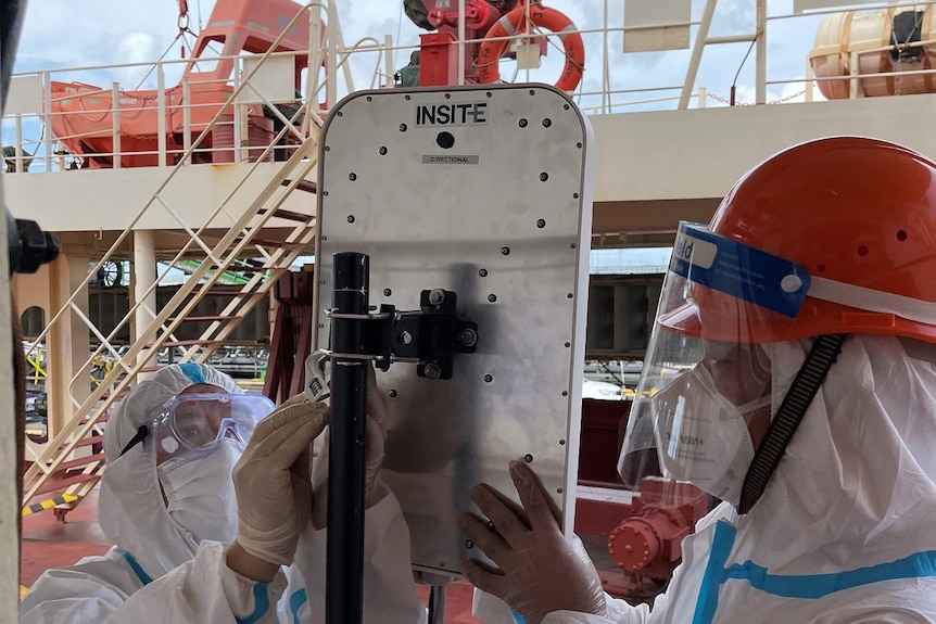 Two people in full PPE using tools to install a large rectangular device labelled INSITE on the deck of a ship.